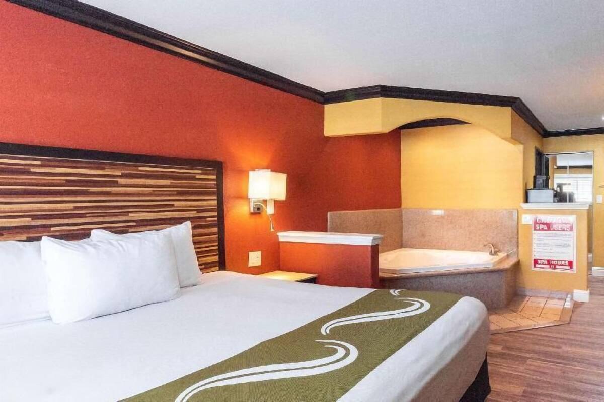 Hotellihuone, jossa on suuri sänky, pieni työpöytä, seinään kiinnitetty valaisin, kylpyamme ja kylpylän aukioloajat ilmoittava kyltti.