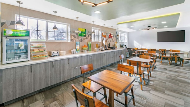 Eine moderne Cafeteria mit Holztischen und -stühlen, einer Theke mit Speisen sowie hängenden Leuchten.