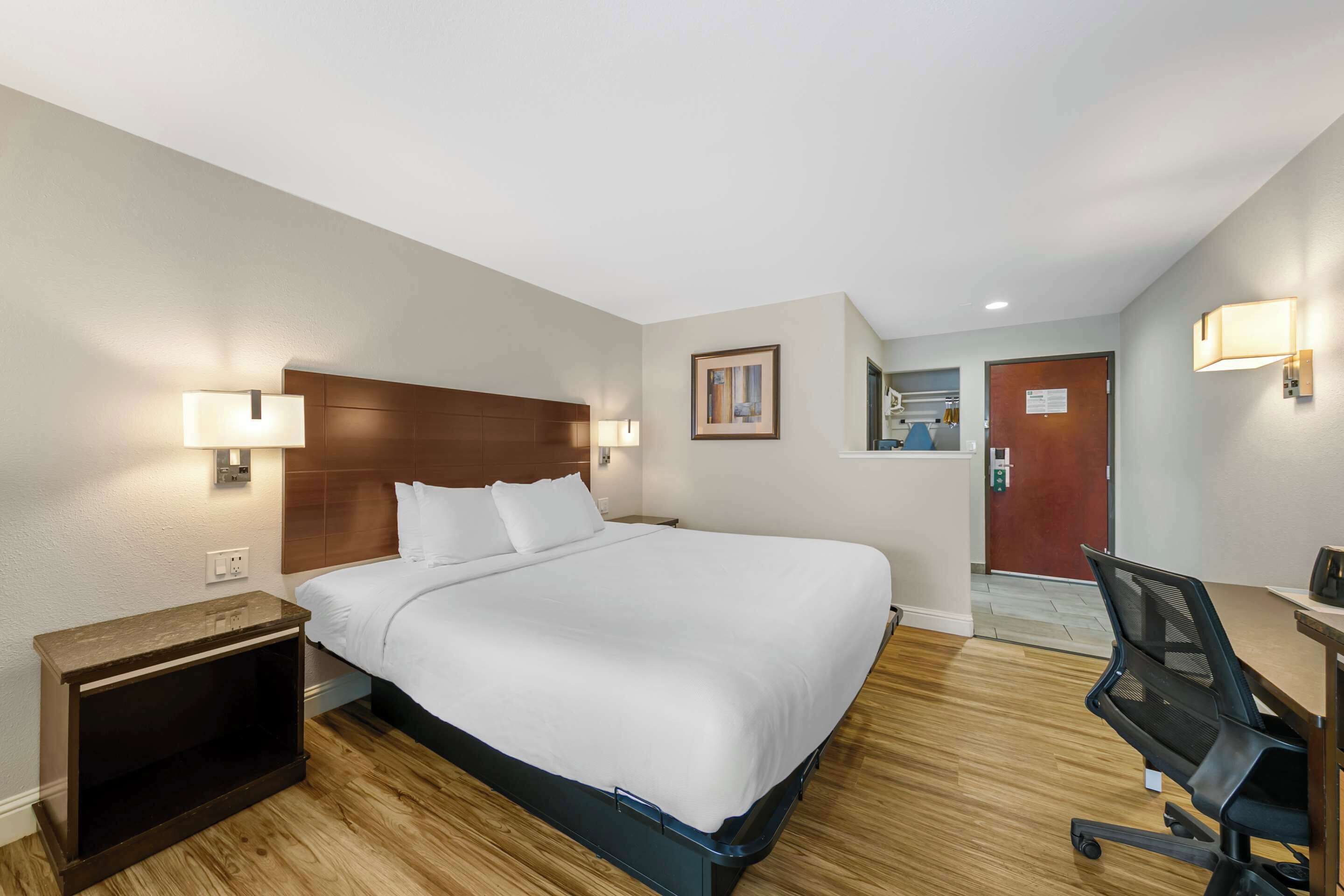 A hotel room with a large bed, a desk with a chair, a nightstand, and a wall-mounted lamp.
