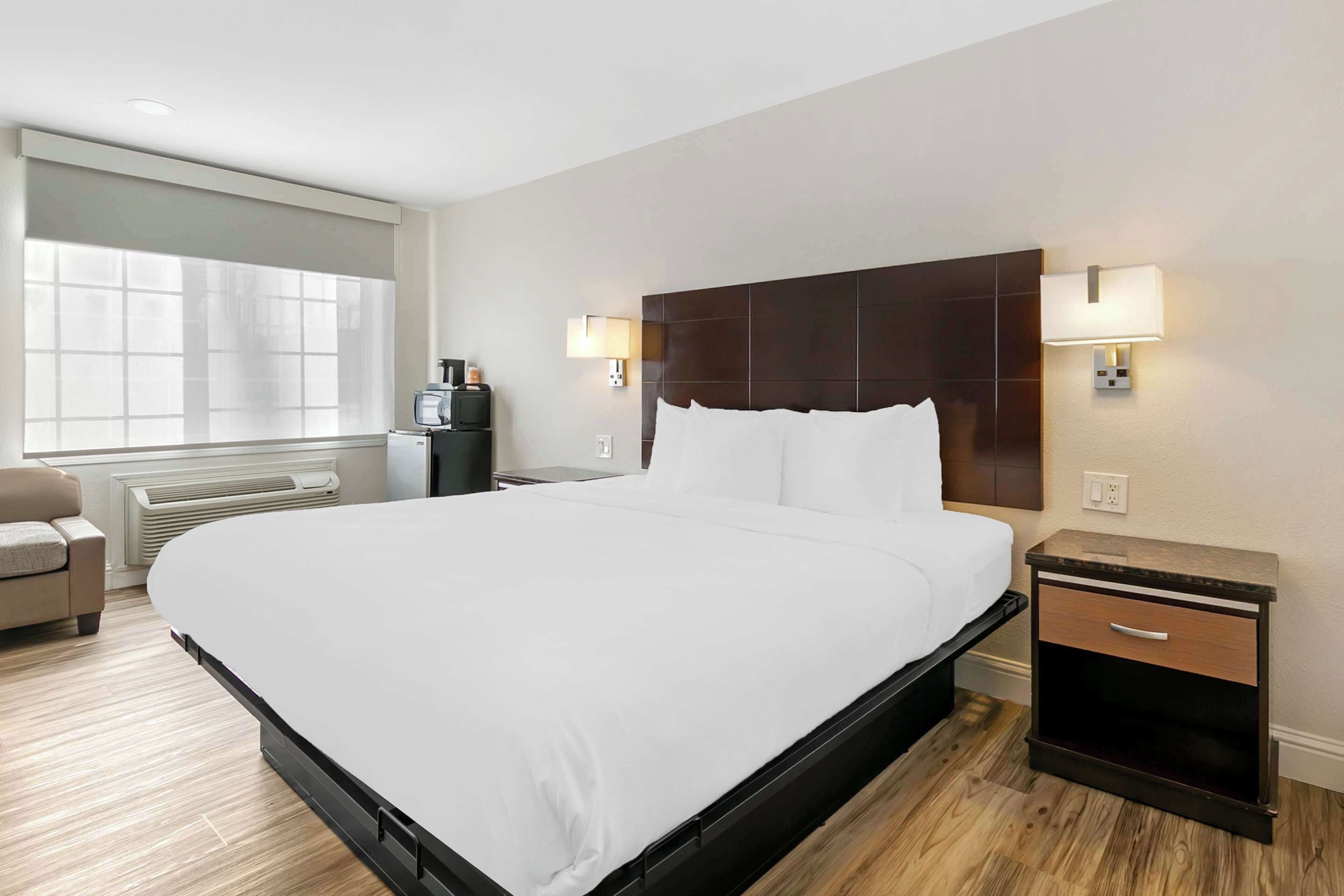 A modern hotel room with a large bed, a bedside table with a lamp, a chair, and a heater.