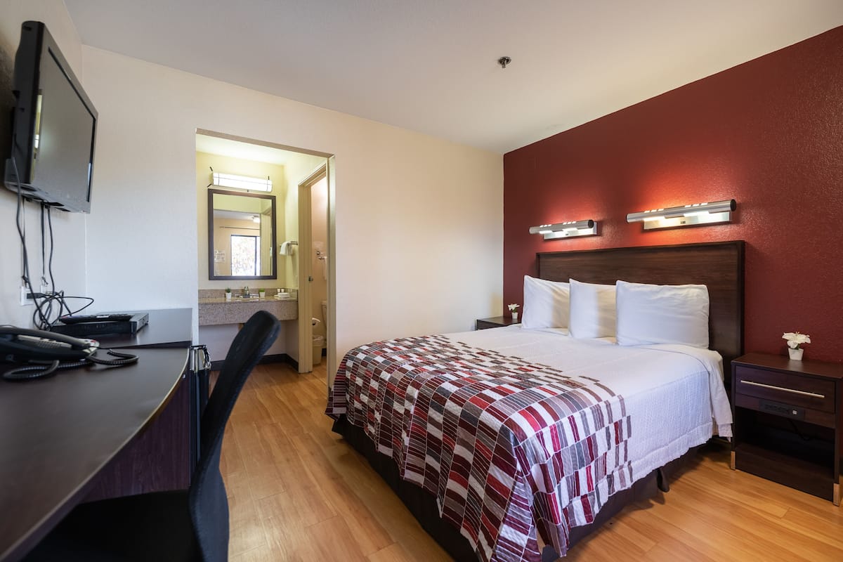 A hotel room with a bed, a desk, a chair, a TV, and a bathroom visible through an open door.