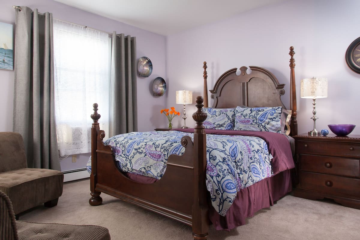 A bedroom with a wooden four-poster bed, a nightstand with a lamp, a chair, and a dresser.