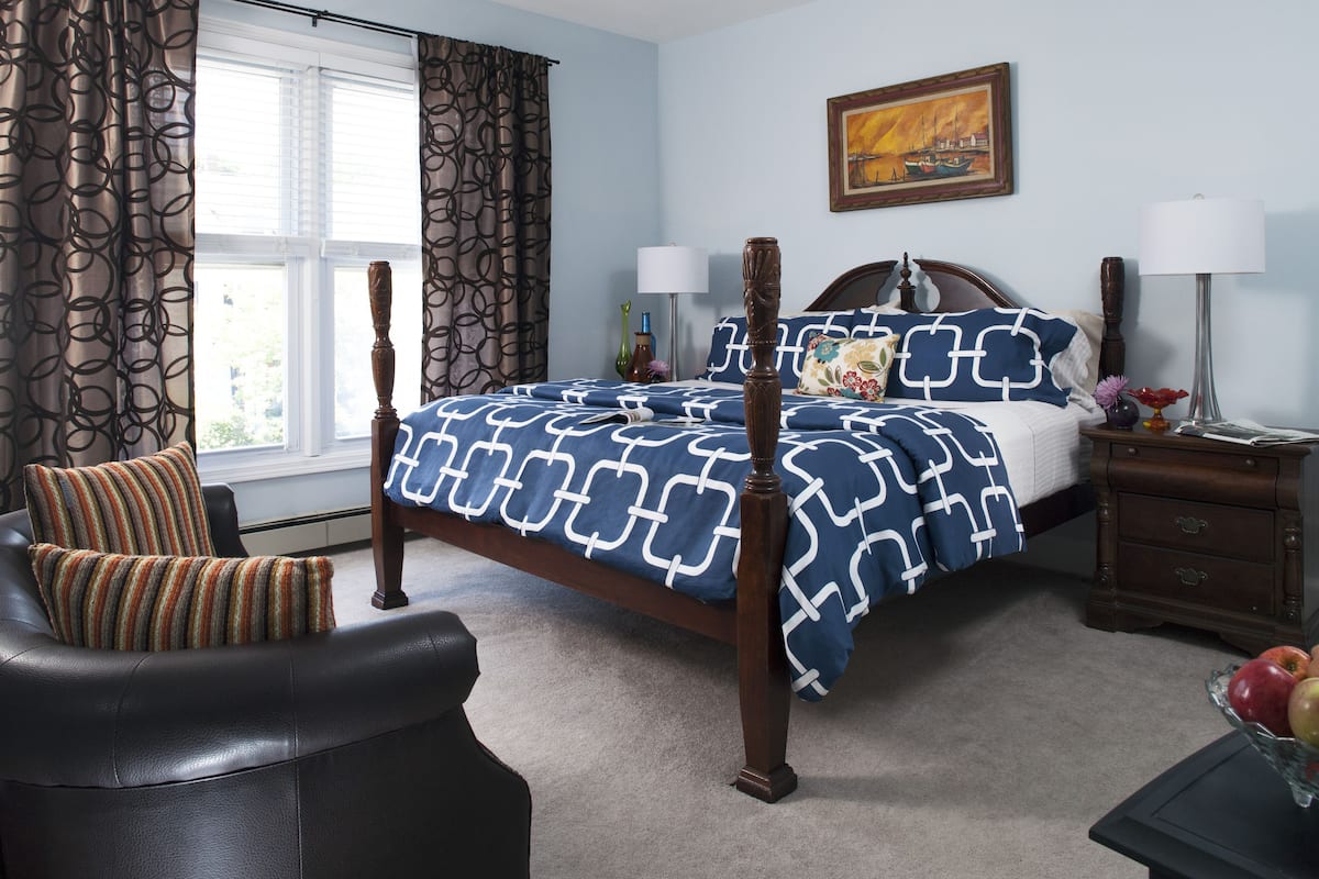 A bedroom with a four-poster bed, a chair, a nightstand, and a window with curtains.