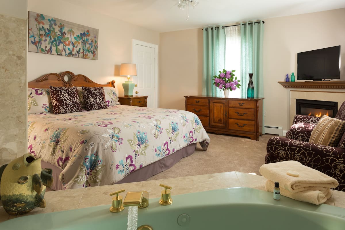 A bedroom with a bed, a vanity with a mirror, a bathtub, a dresser, a television, and a fireplace.