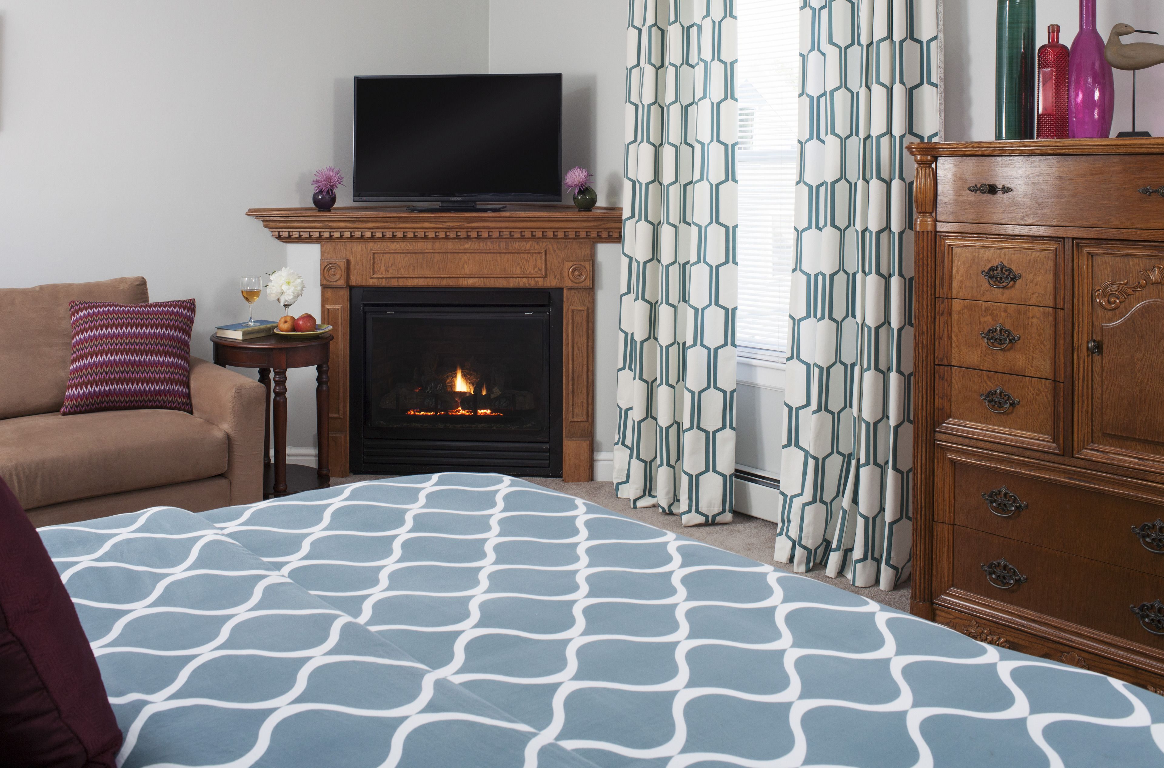 A bedroom with a fireplace, a television, a wooden dresser, and patterned curtains.
