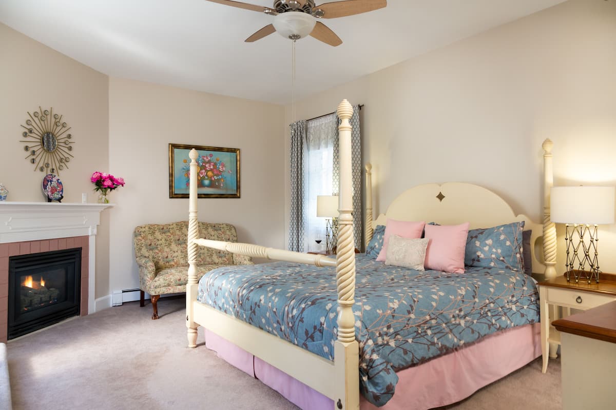 A bedroom with a four-poster bed, a fireplace, a floral armchair, and a nightstand with a lamp.