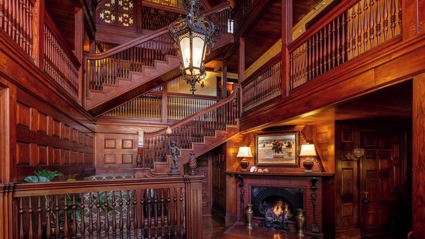 A grand wooden staircase with a chandelier, a fireplace with a painting above it, and a wooden railing. - Ivy Lodge Bed & Breakfast (Newport)