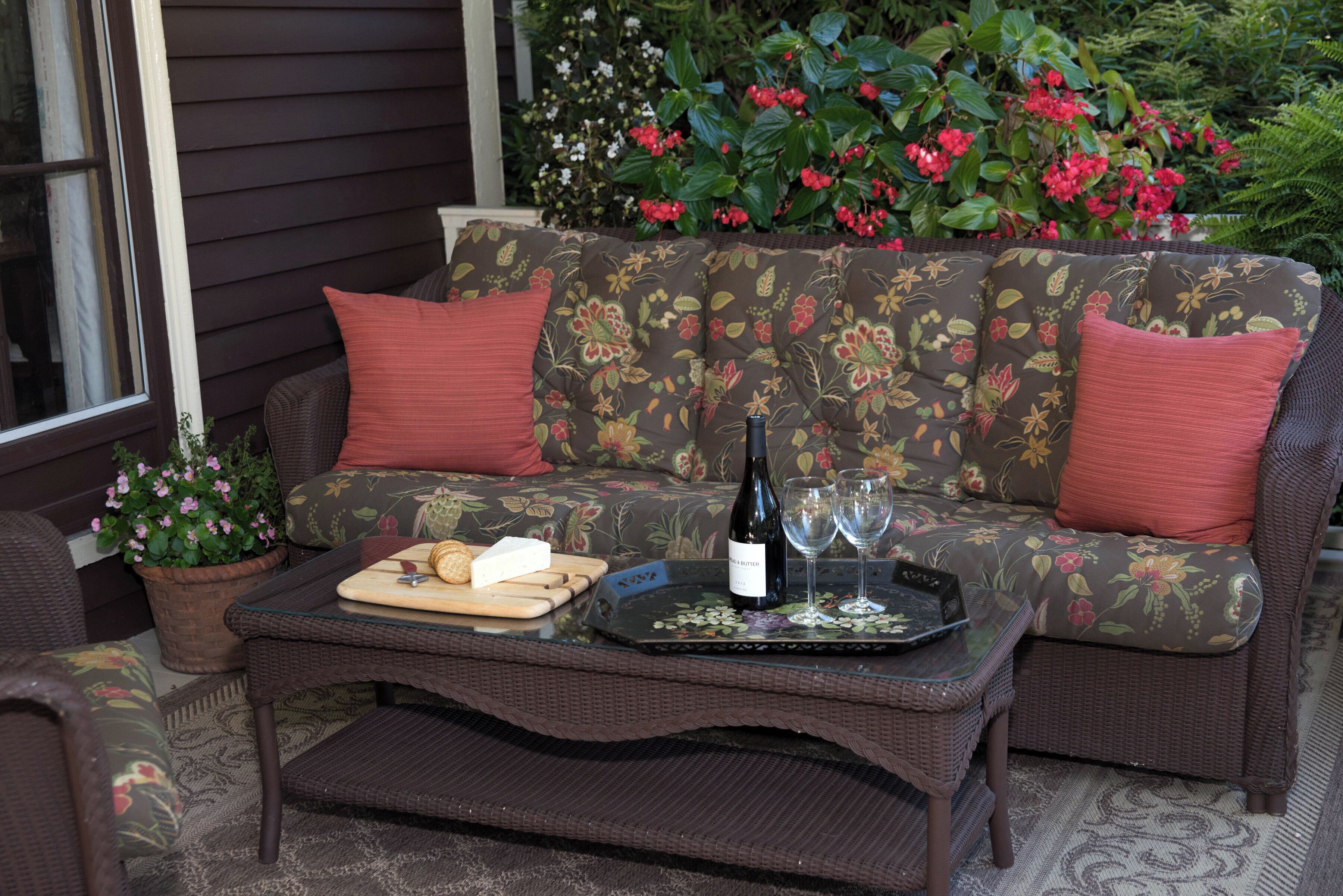 A wicker sofa with floral cushions and red pillows, a glass-top coffee table with a bottle of wine, glasses, and a cheese board.