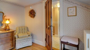 A room with a wooden dresser, a chair, a small table with a blanket, a door leading to another room, and a wall with floral wallpaper. - Melville House (Newport)