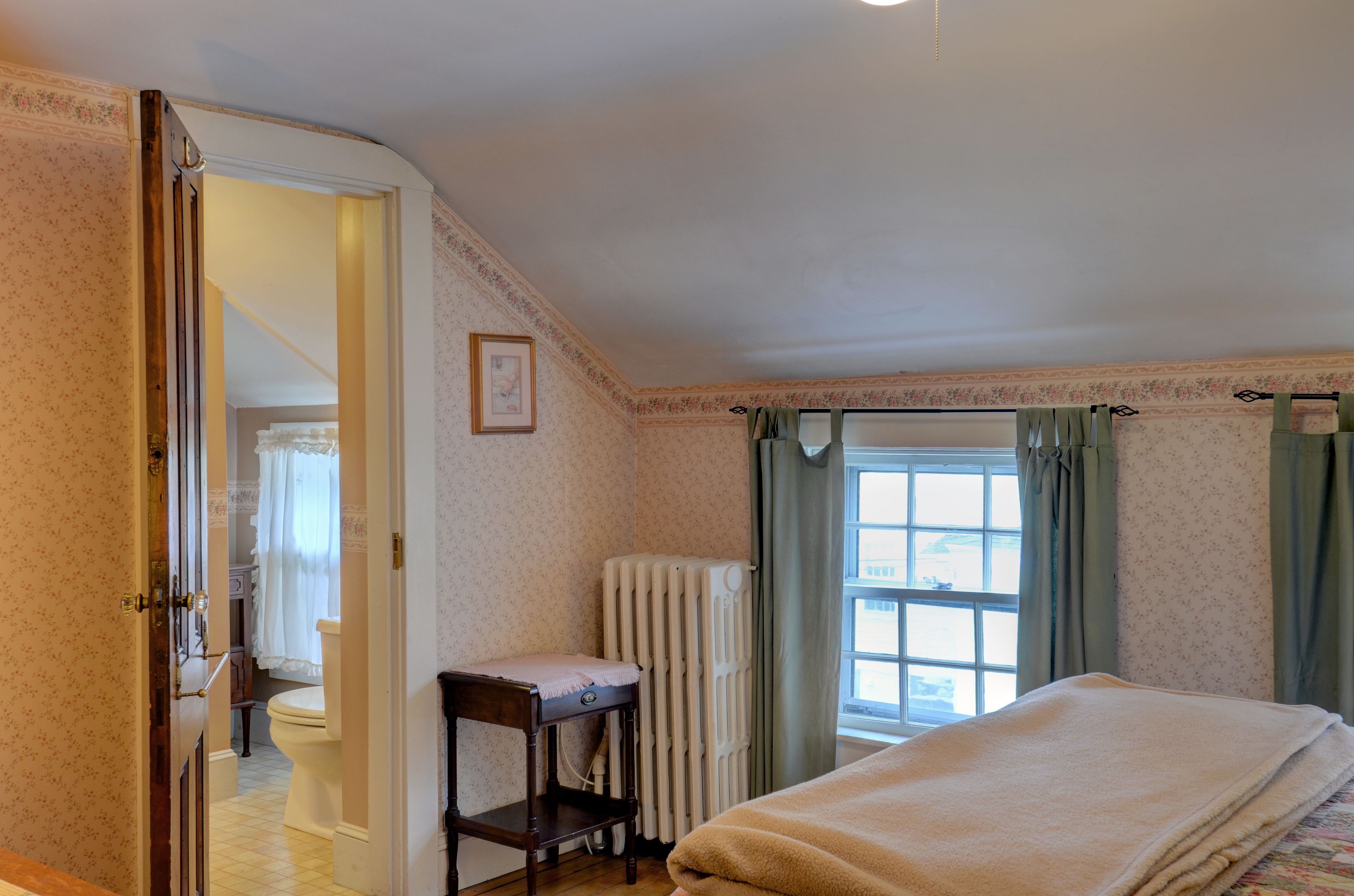 Un dormitorio con cama, un radiador, una ventana con cortinas, una puerta que da al baño y una pequeña mesa con silla.