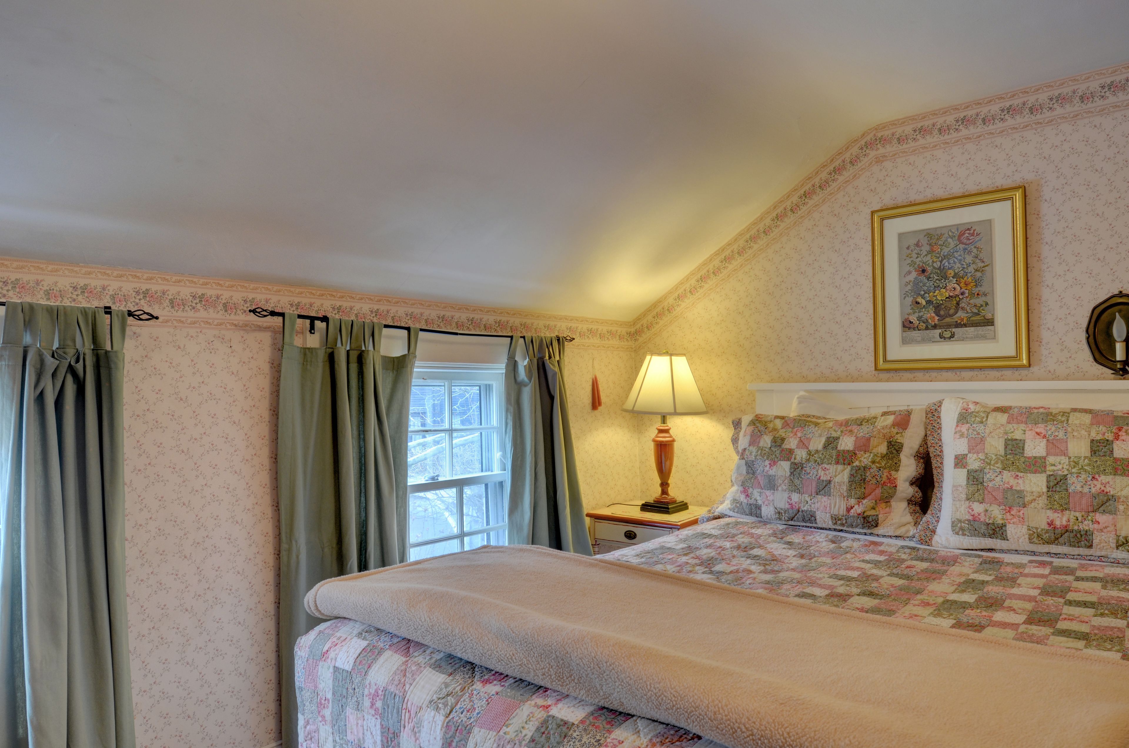A bedroom with a bed, a nightstand, a lamp, a window with curtains, and a floral-patterned wall.