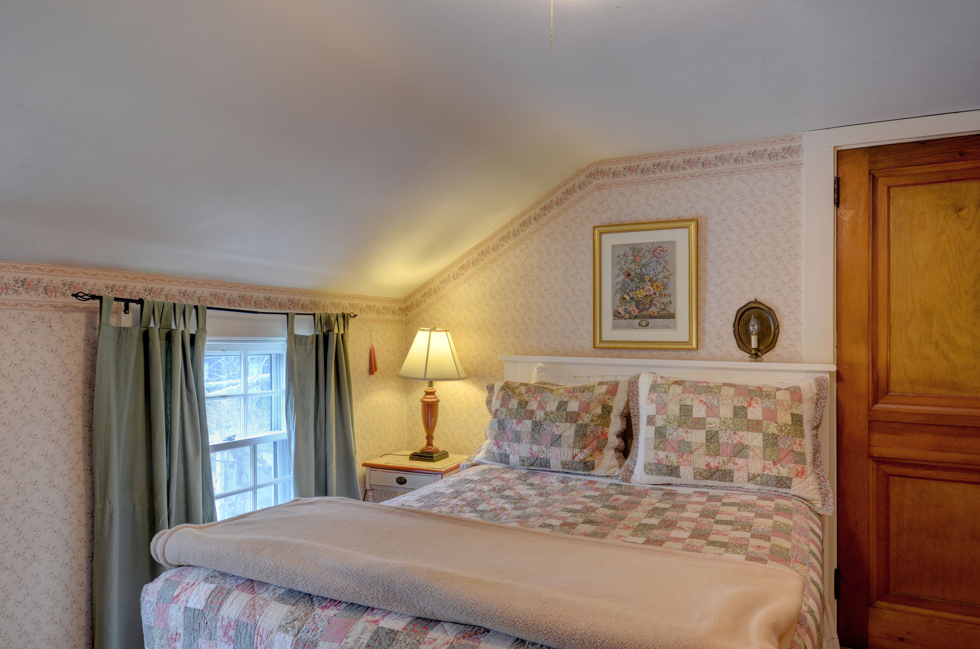 A bedroom with a bed, a nightstand, a lamp, a framed picture, and a wooden door.