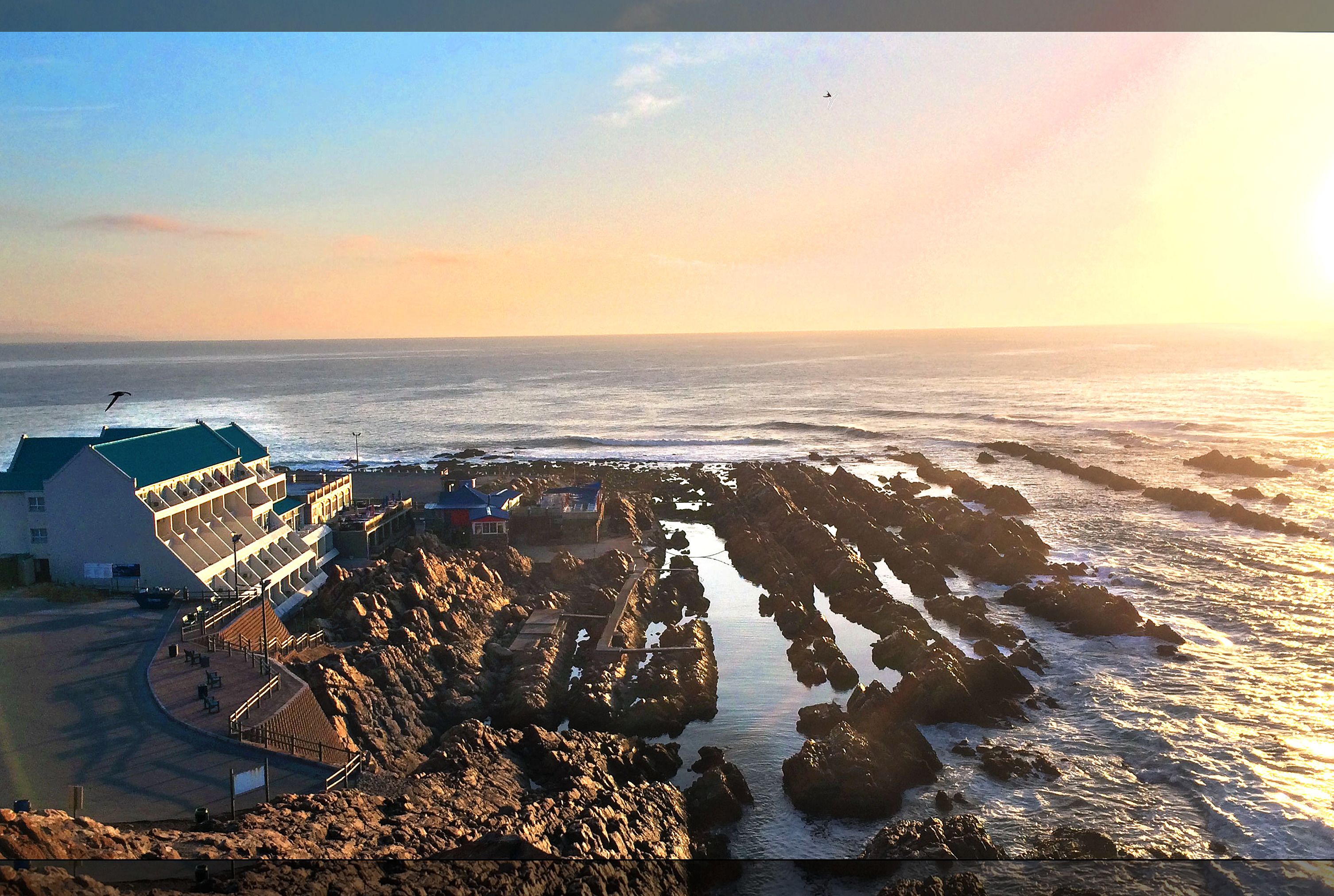 Un edificio frente al mar con techo curvo, un camino que lleva al océano y terreno rocoso.
