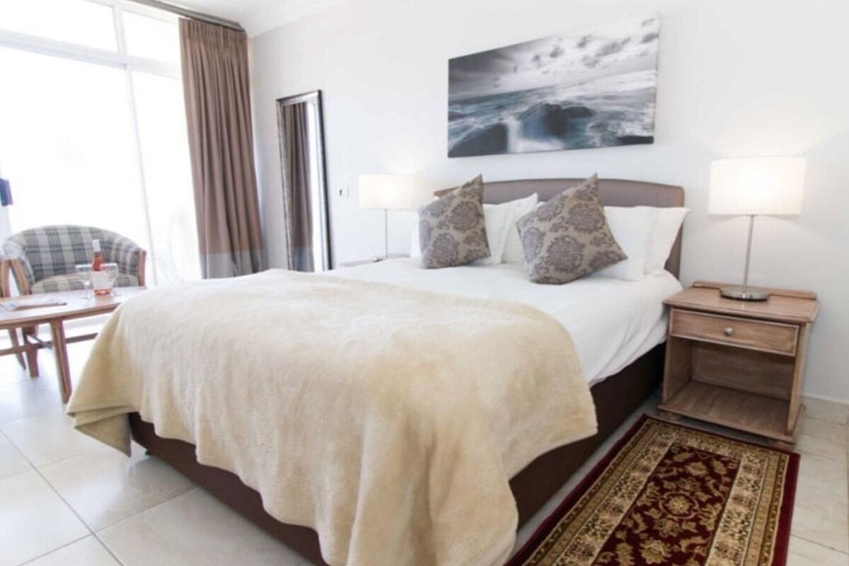 A hotel room with a large bed, a bedside table with a lamp, a chair, and a view of the sea through the window.