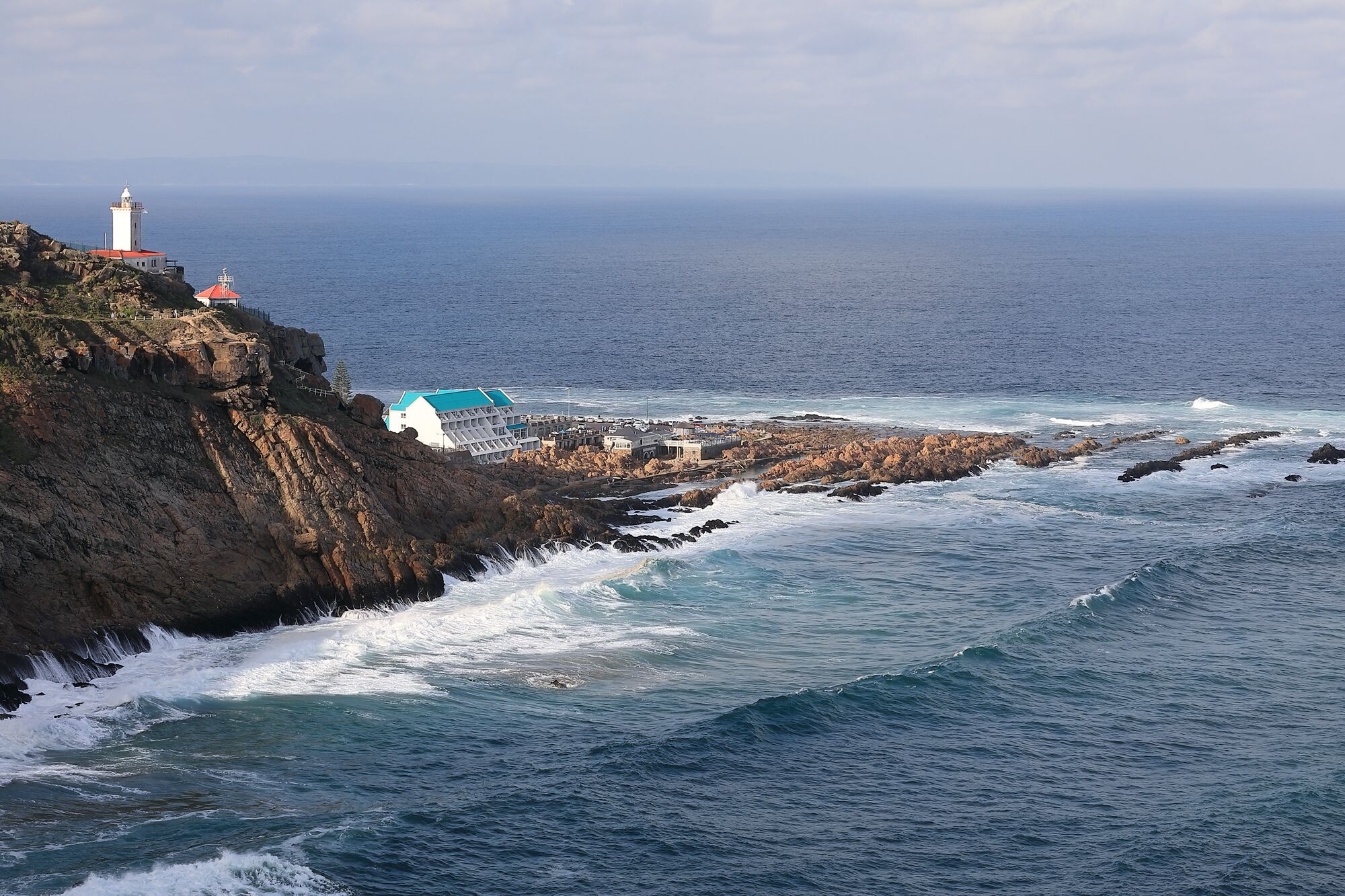 Un edificio costero con un faro, acantilados rocosos y olas rompiendo en la orilla.