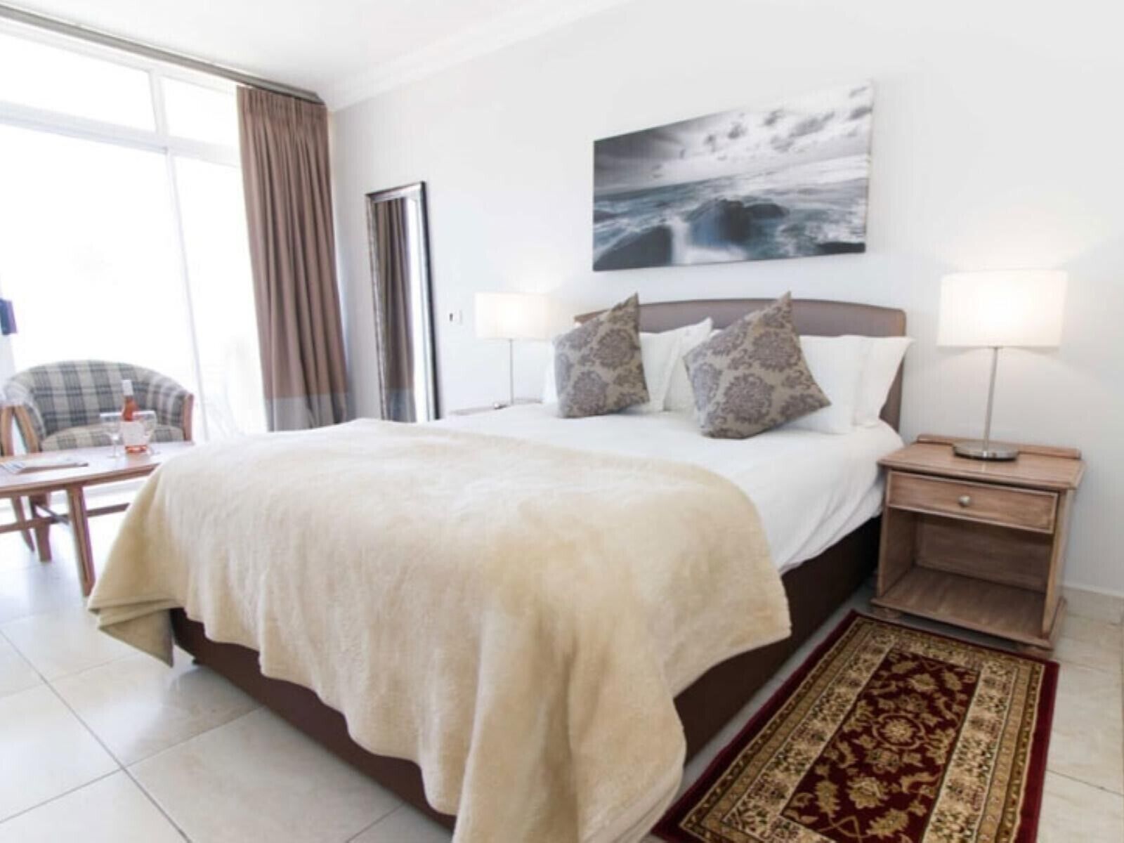 A hotel room with a large bed, a bedside table with a lamp, a chair, and a view of the sea through the window.