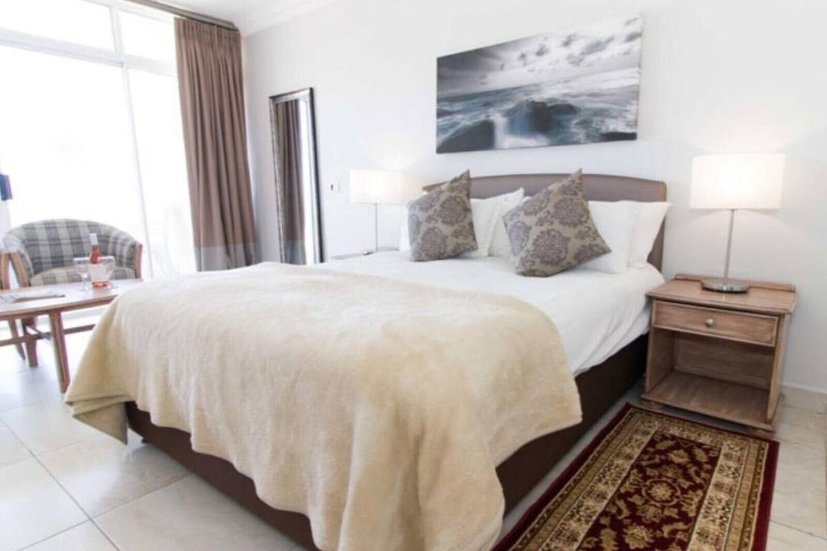 A hotel room with a large bed, a bedside table with a lamp, a chair, and a view of the sea through the window.