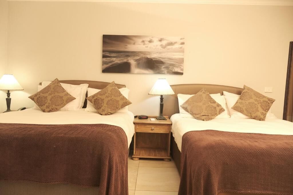 A hotel room with two beds, each with a brown bedspread, a nightstand with a lamp, and a wall-mounted picture of a seascape.