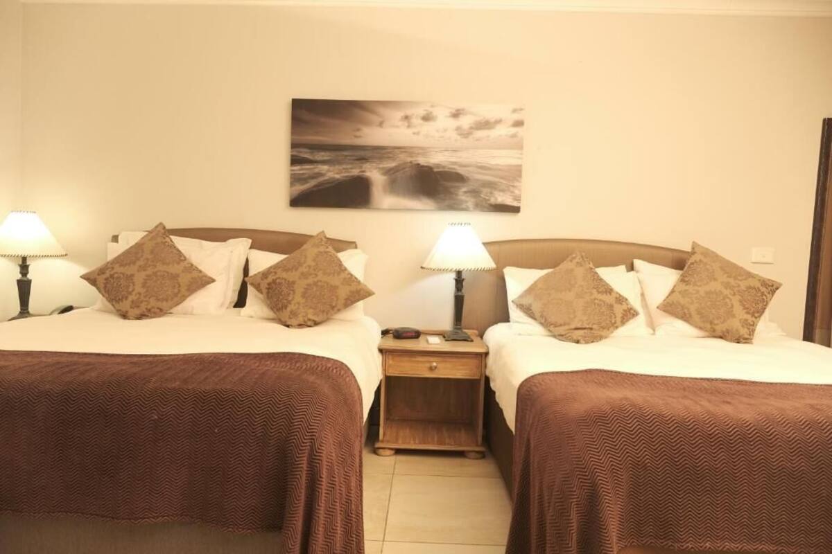 A hotel room with two beds, each with a brown bedspread, a nightstand with a lamp, and a wall-mounted picture of a seascape.
