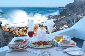 A table with a meal, glasses, and a view of the ocean and rocky coastline. - The Point Hotel & Spa (Mossel Bay)