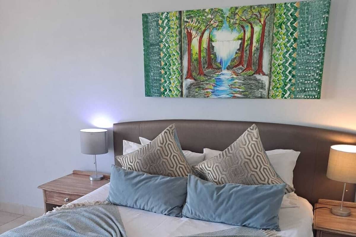 A neatly made bed with patterned pillows, a blanket, and a rolled towel. There are two bedside tables with lamps. A painting of a forest scene is on the wall above the bed.