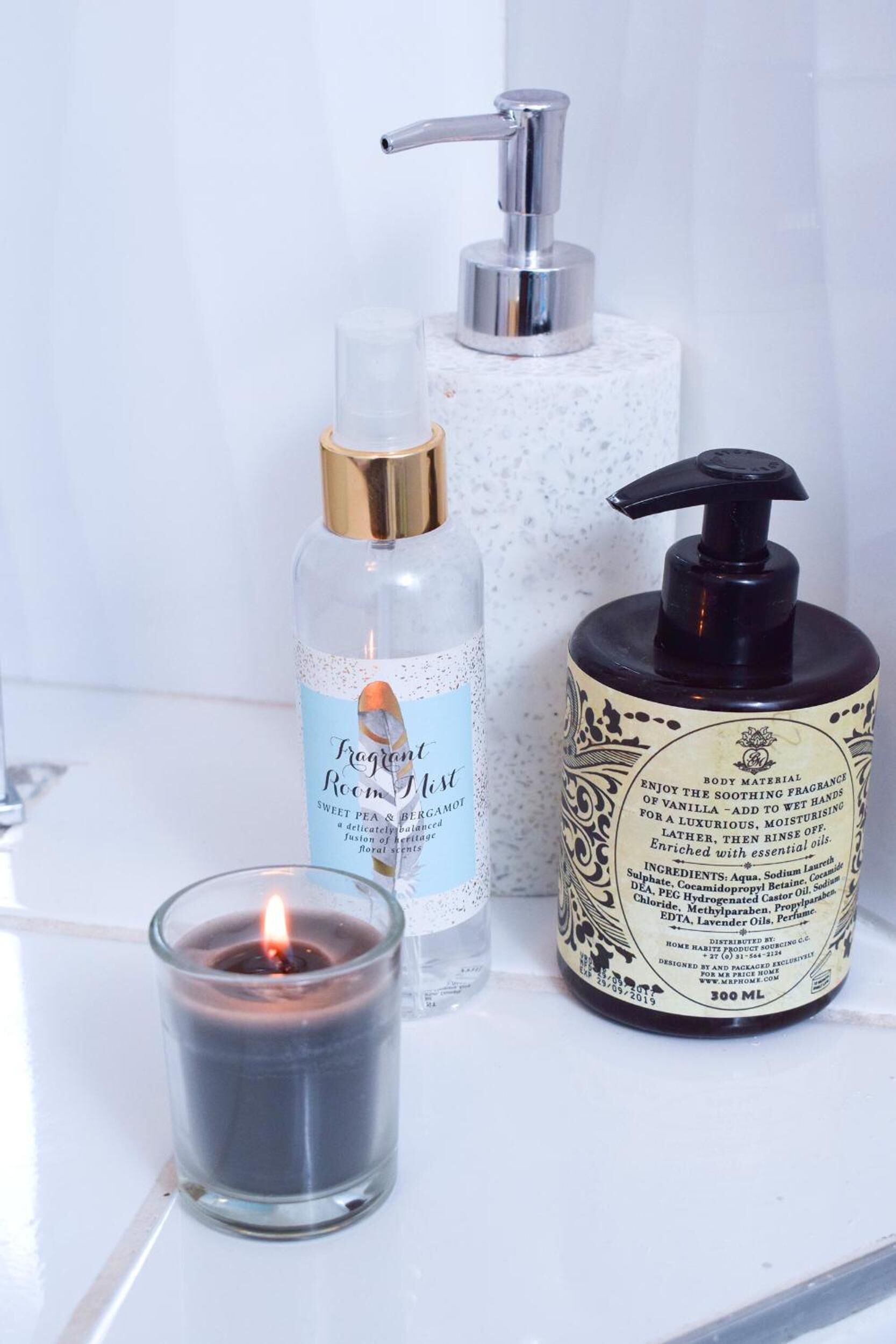 Une salle de bain avec un lavabo blanc, une bougie en verre et deux flacons de produits de soins pour le corps.