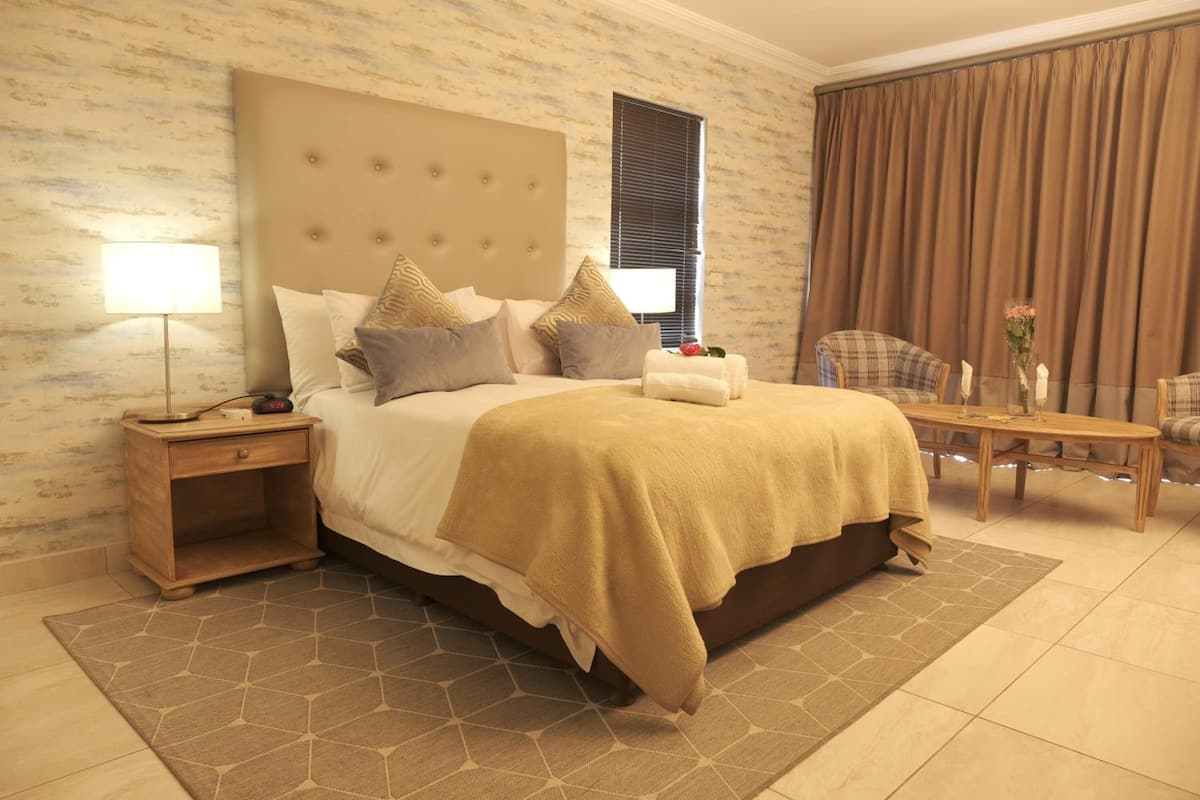 A hotel room with a large bed, a bedside table with a lamp, a chair, a small table with a vase, and a window with curtains.
