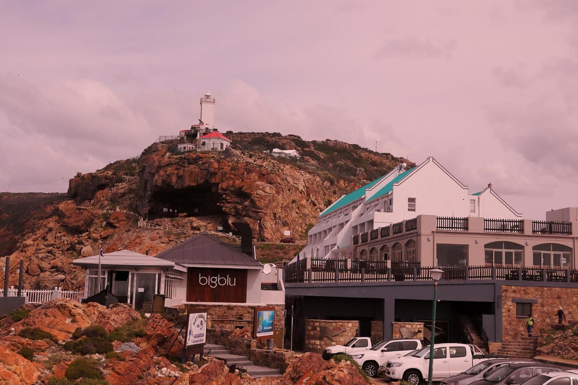 A coastal hotel with a prominent lighthouse on a rocky hill, a modern building with a blue roof, and a sign for 'bigblu' restaurant.