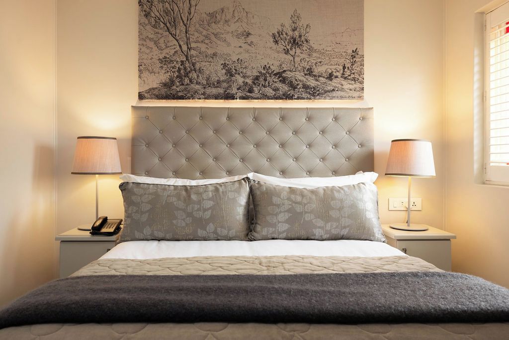 A neatly made bed with a tufted headboard, two bedside lamps, a telephone, and a landscape painting above the headboard.