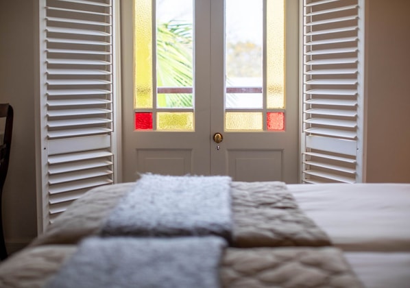 Un dormitorio con una cama, una puerta con paneles de vidrio esmerilado y contraventanas blancas en las ventanas.