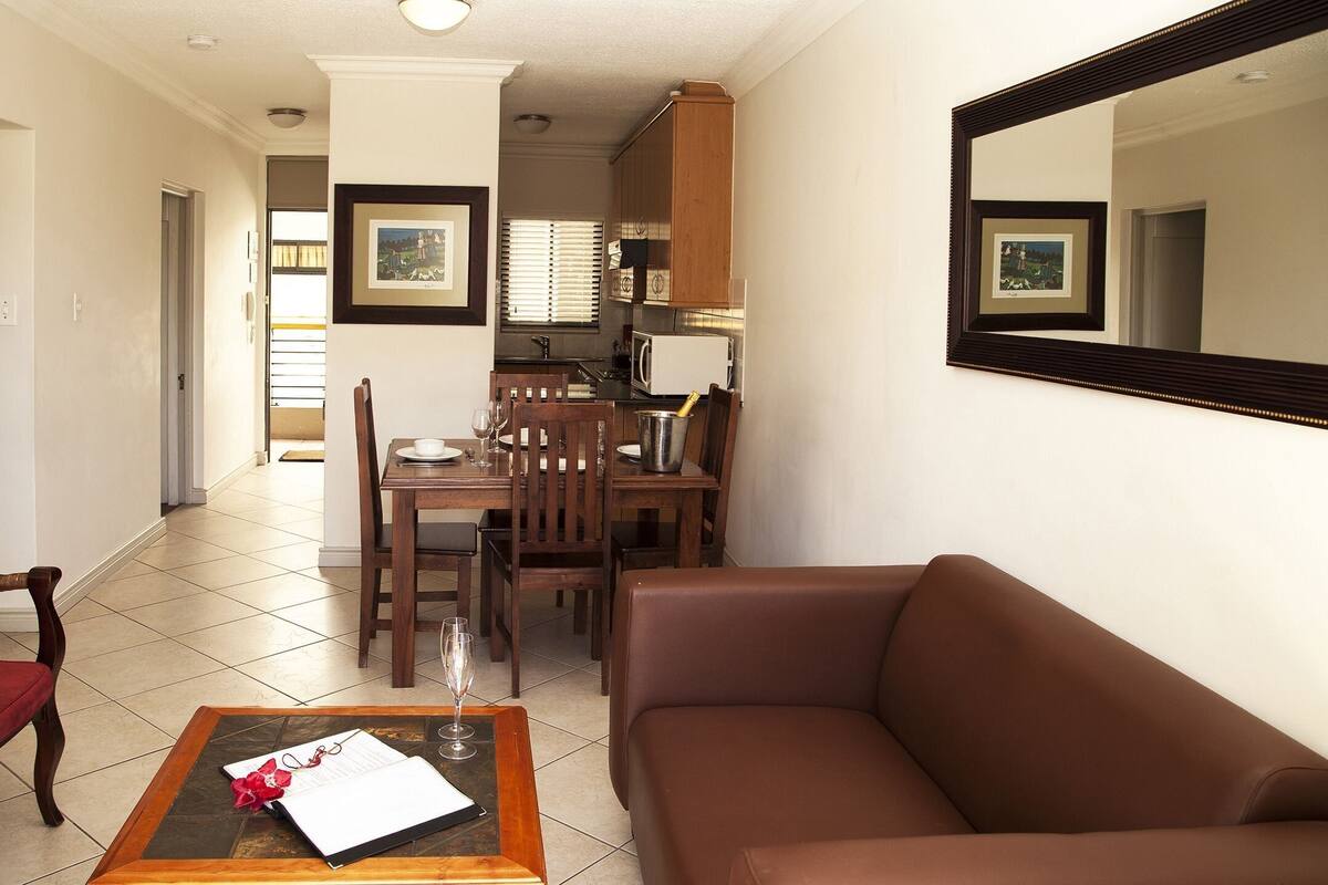A living room with a brown sofa, a dining table with chairs, a kitchen area, and a hallway leading to a bathroom.