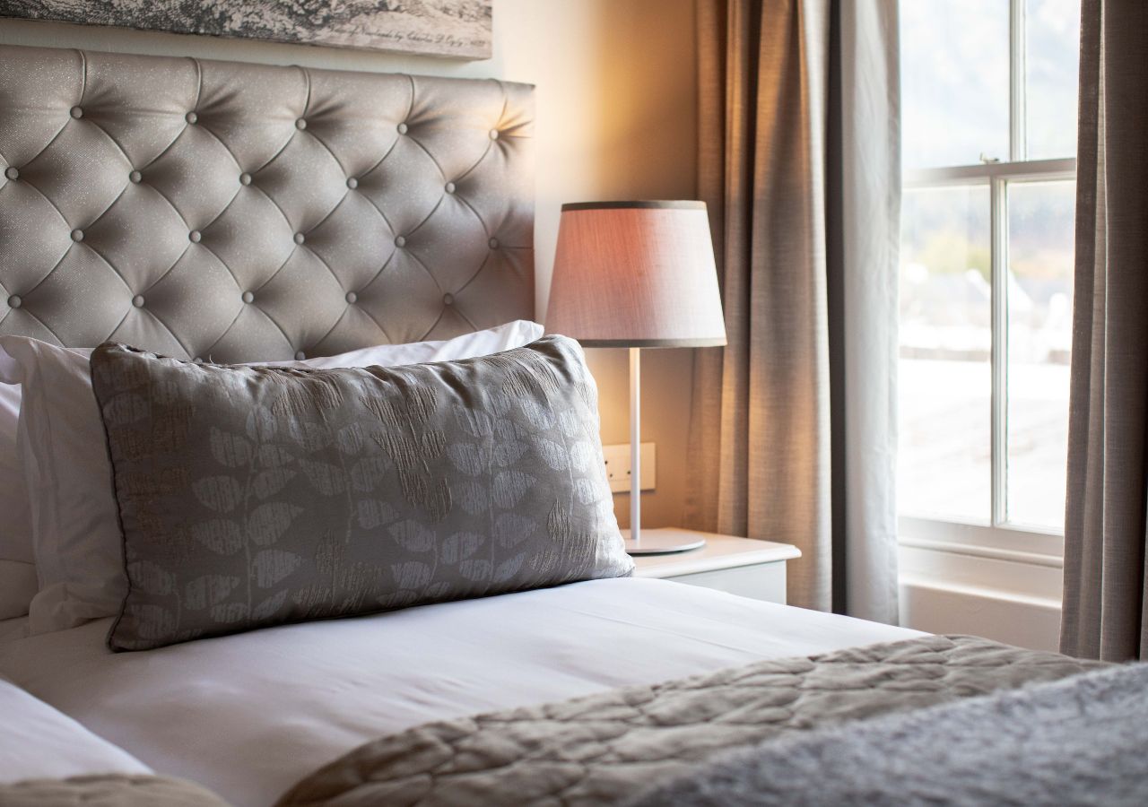 A hotel room with a tufted headboard, a bedside lamp, and a view through a window.
