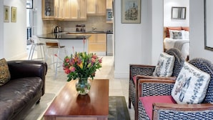 Una sala moderna con un sofá de cuero, sillones estampados y una mesa de centro. Se ve en el fondo una cocina con gabinetes de madera y un comedor con taburetes de bar.