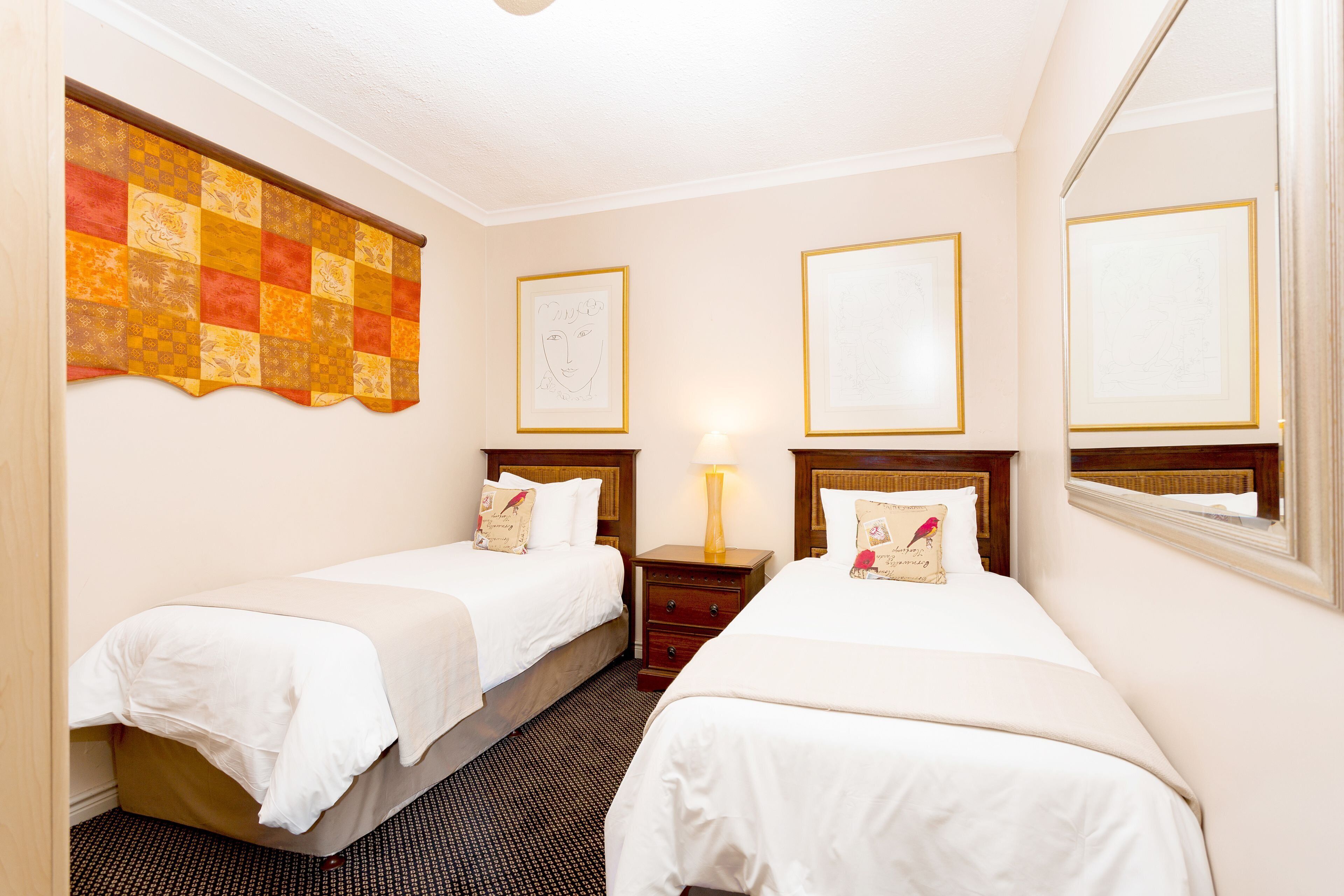 Une chambre d’hôtel avec deux lits, un rideau à carreaux, des œuvres d’art encadrées, une table de chevet avec une lampe et une fenêtre donnant sur un paysage.