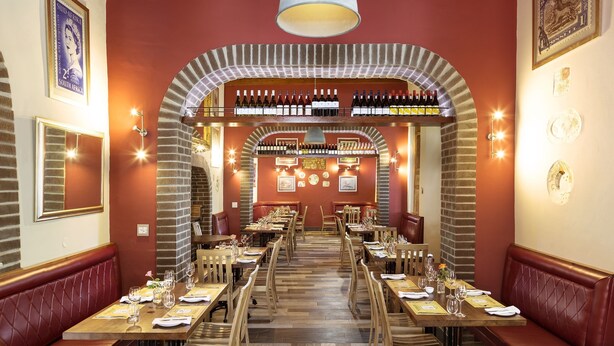 Un restaurante con una larga mesa de madera y asientos tapizados en cuero rojo, arcos de ladrillo y botellas de vino en estanterías.