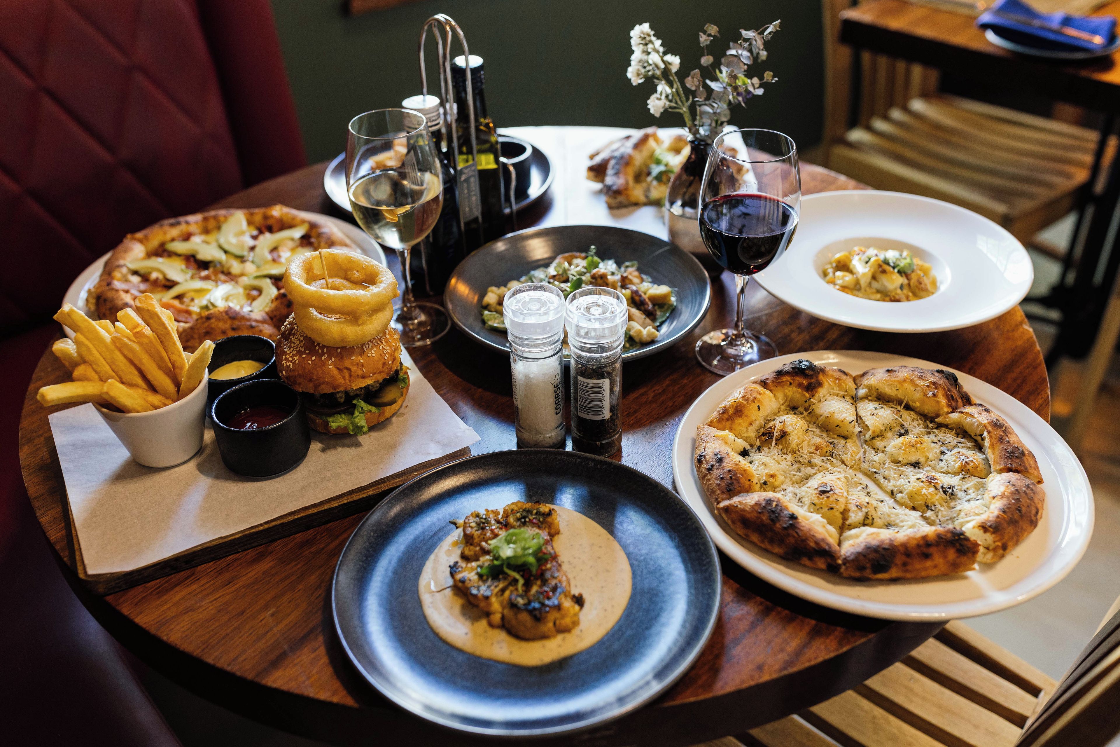 Una mesa de comedor con diferentes platos, incluyendo pizza, papas fritas y una ensalada, además de salsas y bebidas.