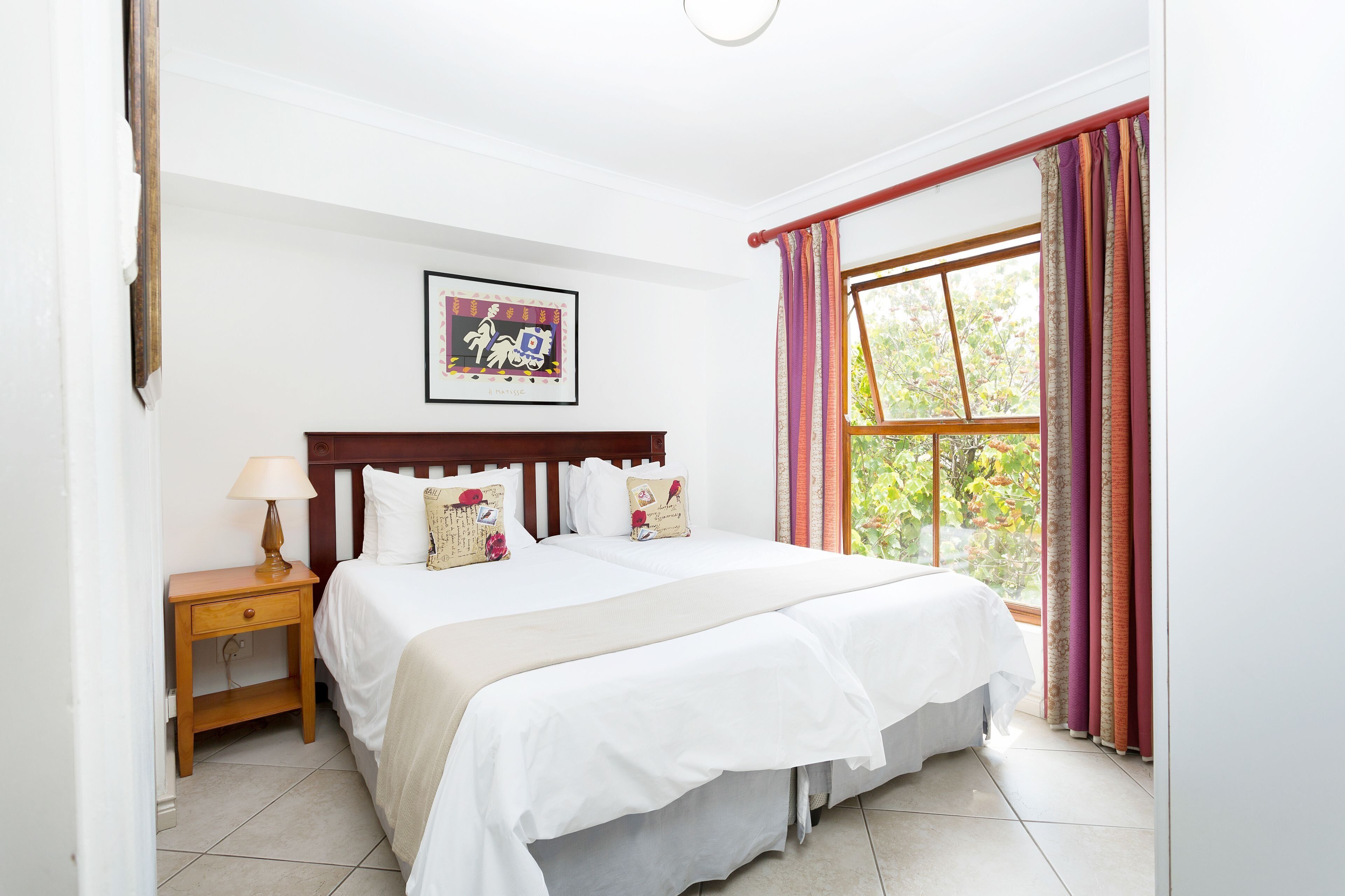 A bedroom with a bed, a nightstand, a lamp, a framed picture, and a window with curtains.
