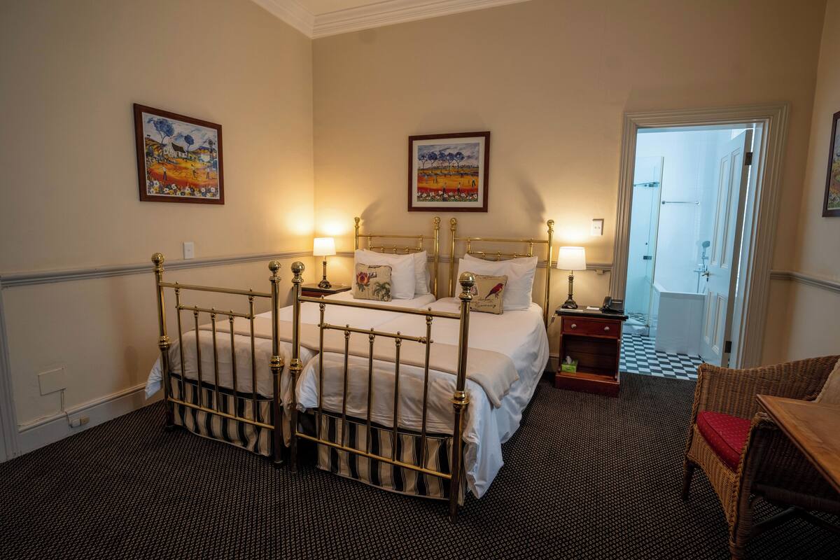 A hotel room with a four-poster bed, two bedside tables with lamps, a wicker chair, and two framed artworks on the walls.