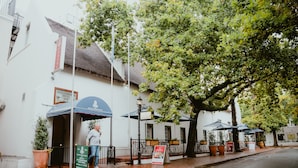 Un hotel con fachada tradicional, un área de estar al aire libre techada y un árbol que proporciona sombra.