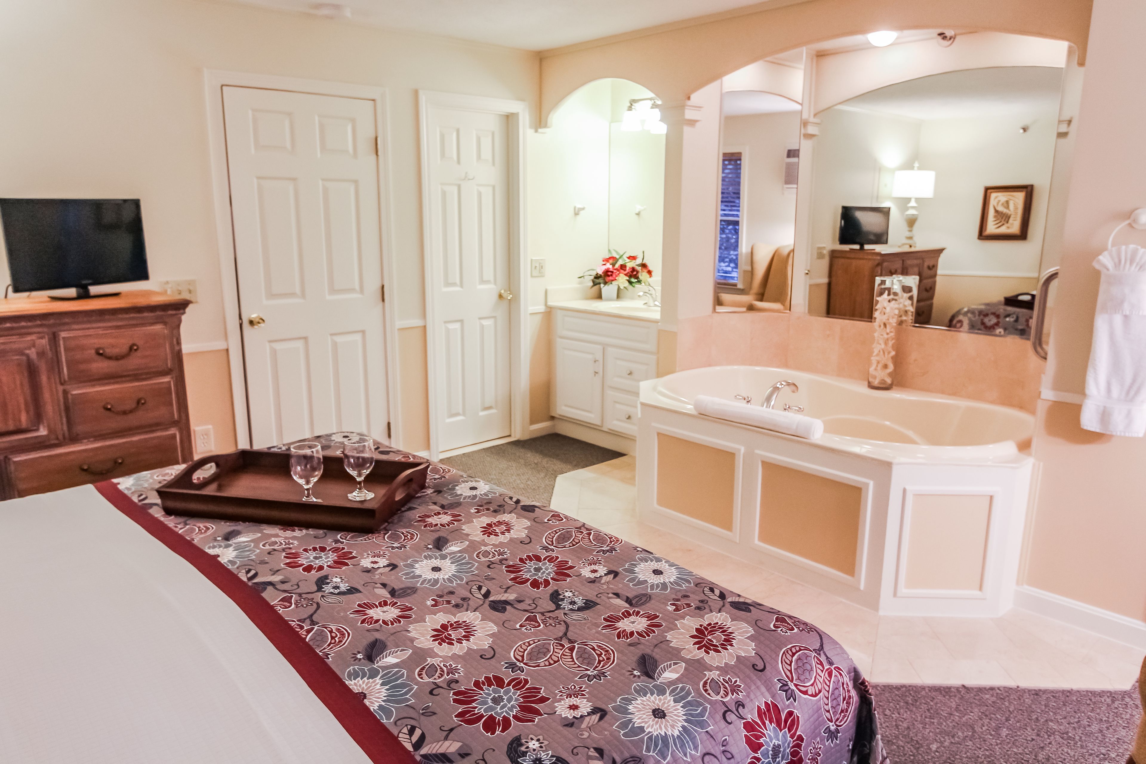 A bedroom with a bed, a dresser, a vanity with a mirror, a bathtub, and a television.