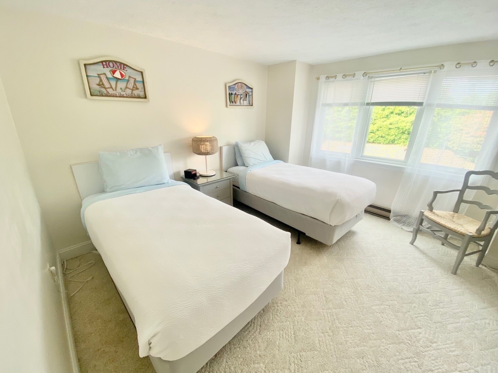 A bedroom with two single beds, a nightstand with a lamp, a chair, and a window with sheer curtains.