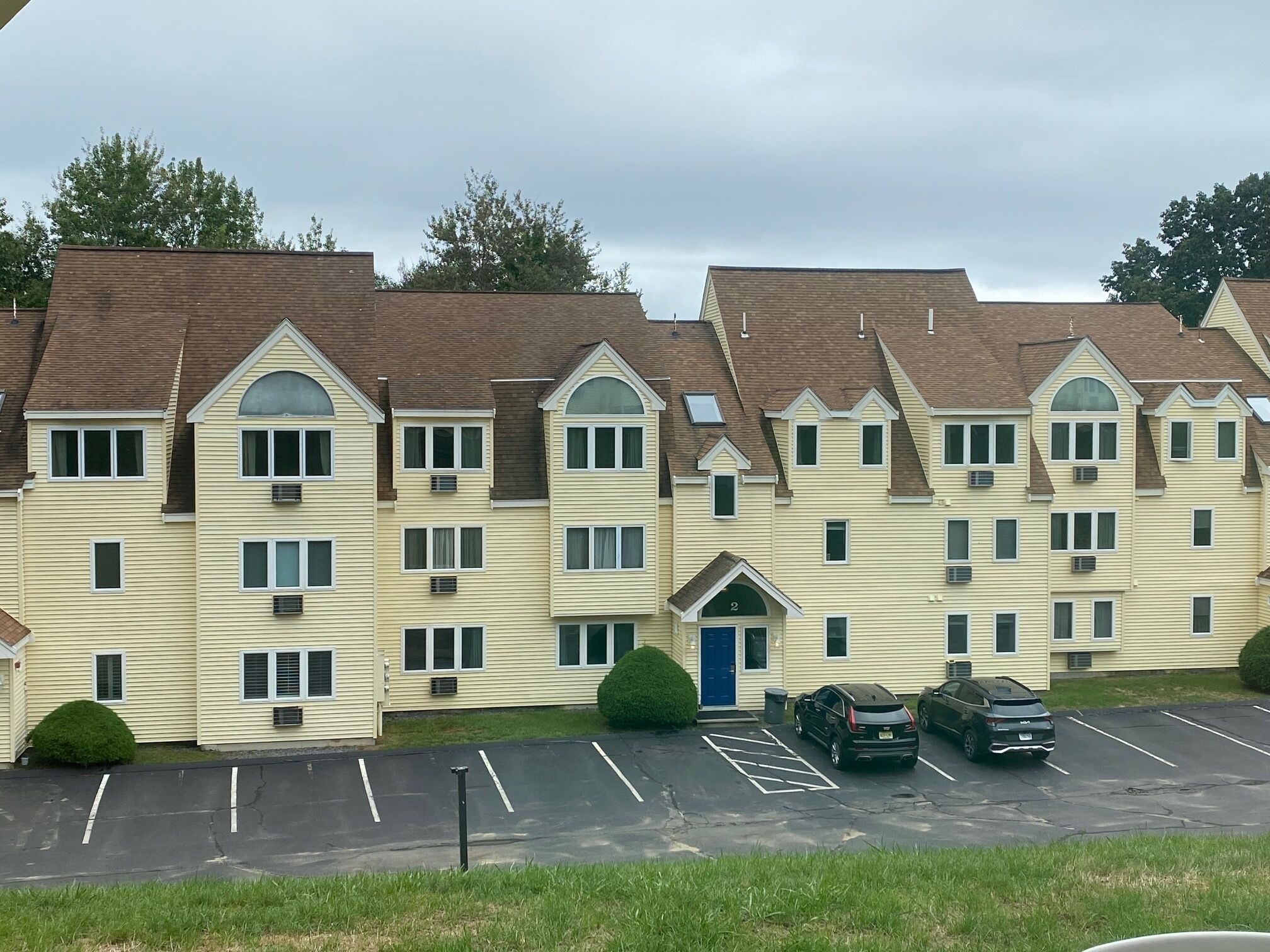 Een meerhoog hotel met een uniforme architectuur, met een centrale ingang met een blauwe deur, meerdere ramen en een parkeerplaats met verschillende auto's.