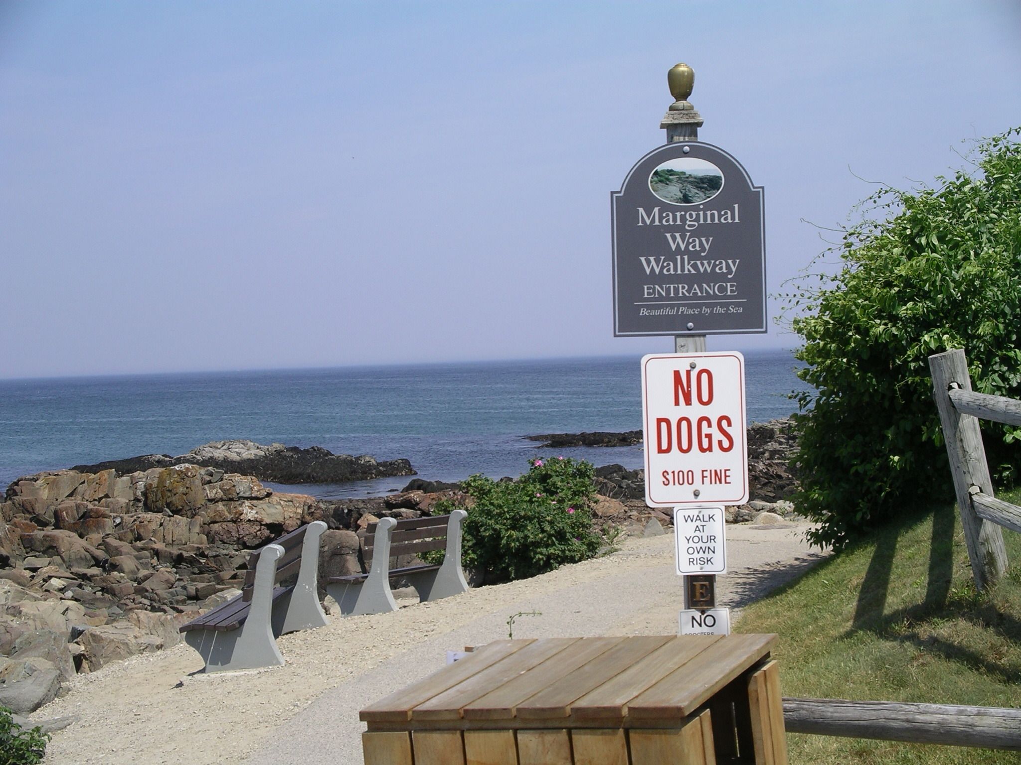 Señal de entrada a un paseo marítimo en la playa con un letrero que indica 'No se permiten perros' y un aviso de multa de 100 dólares por perros.