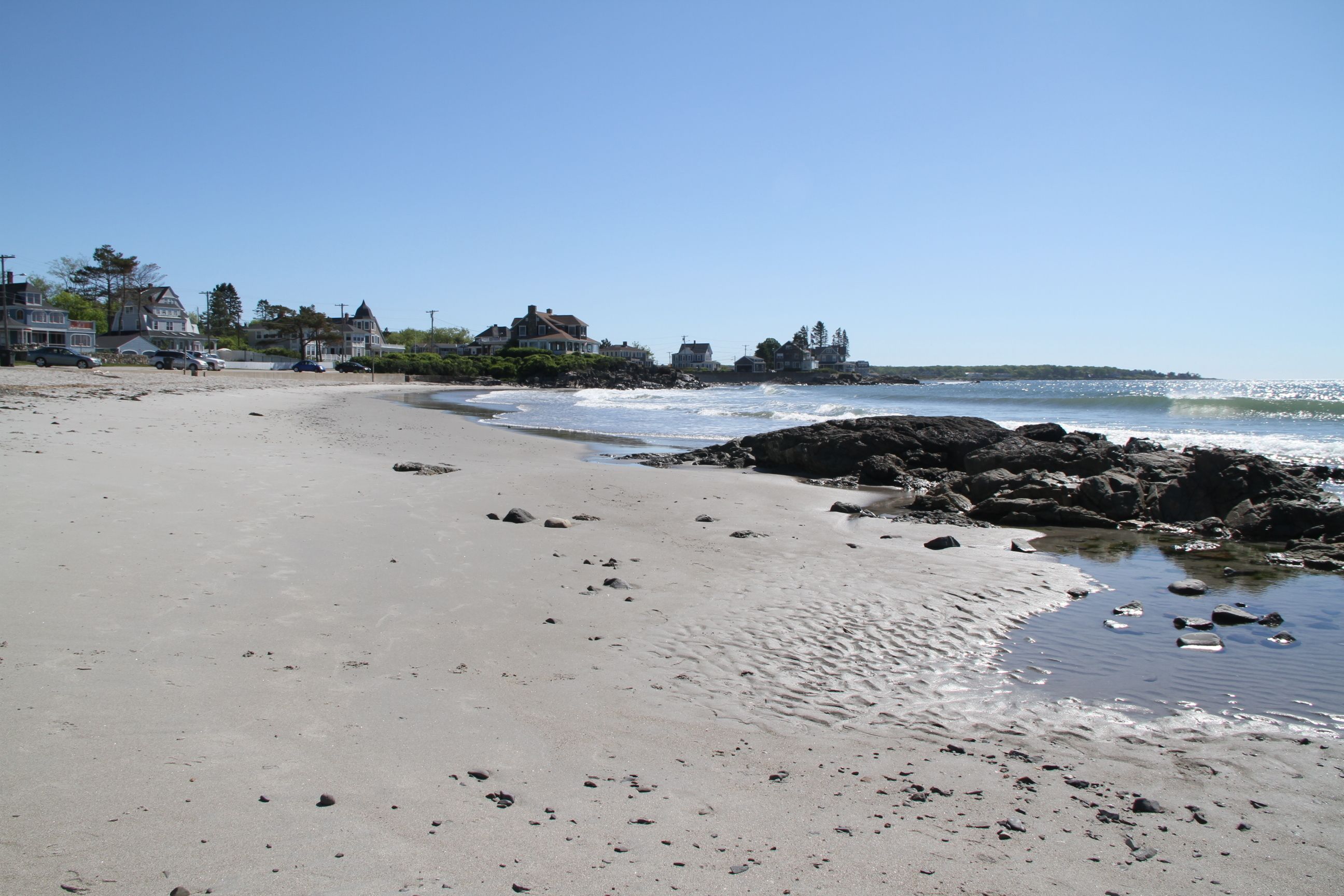Une plage de sable parsemée de rochers, un ciel bleu dégagé et des maisons en arrière-plan.