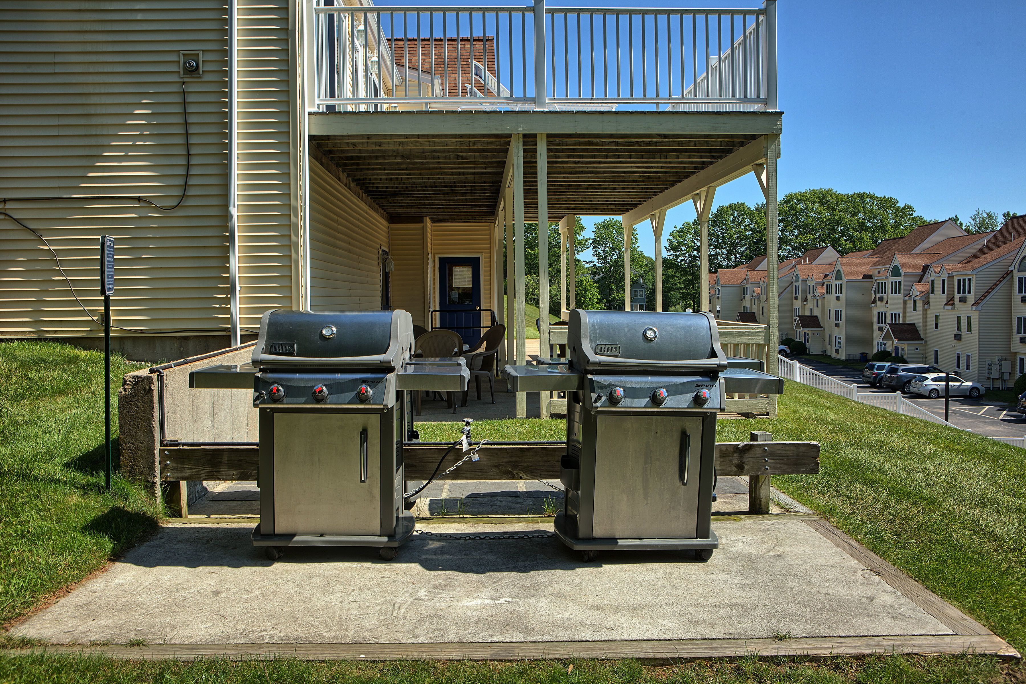 A covered outdoor area with two barbecue grills, a wooden bench, and a door leading to a building with a balcony.
