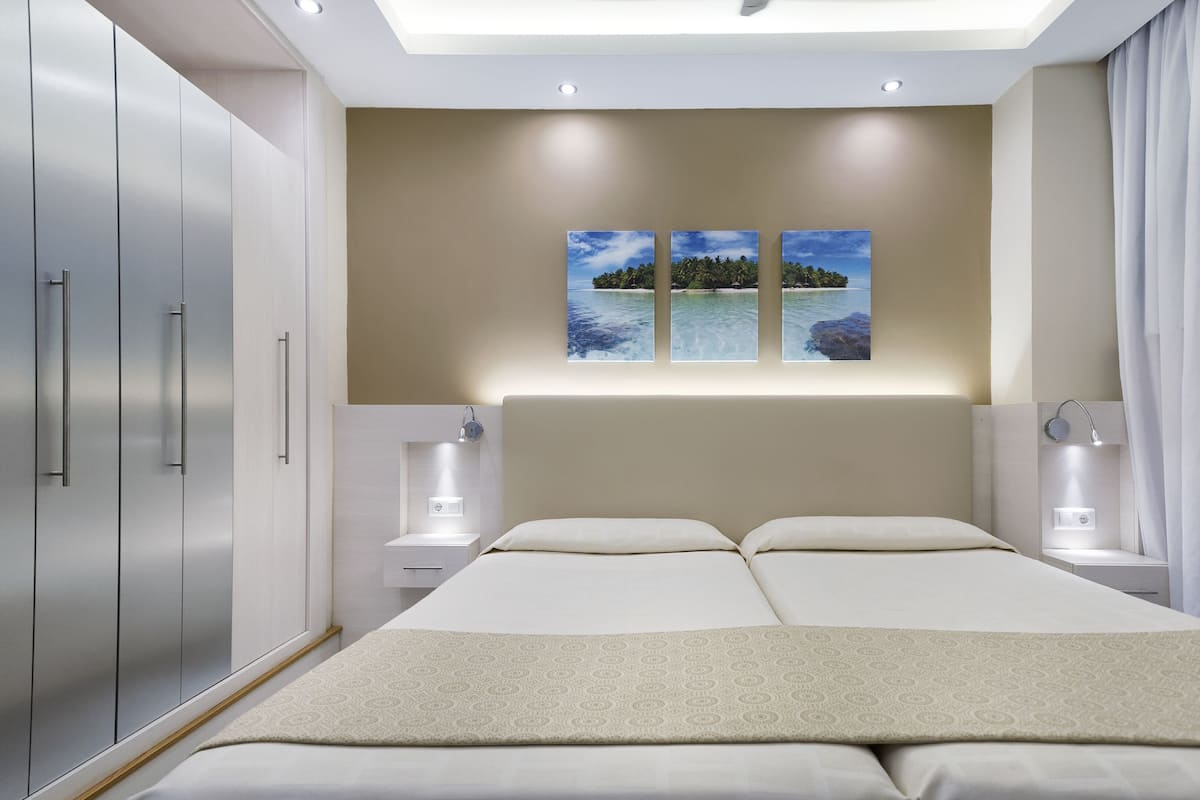 Ein modernes Schlafzimmer mit einem großen Bett, Nachttischen und einem Kleiderschrank. Zwei Bilderrahmen sind an der Wand angebracht und über dem Bett gibt es ein Oberlicht.
