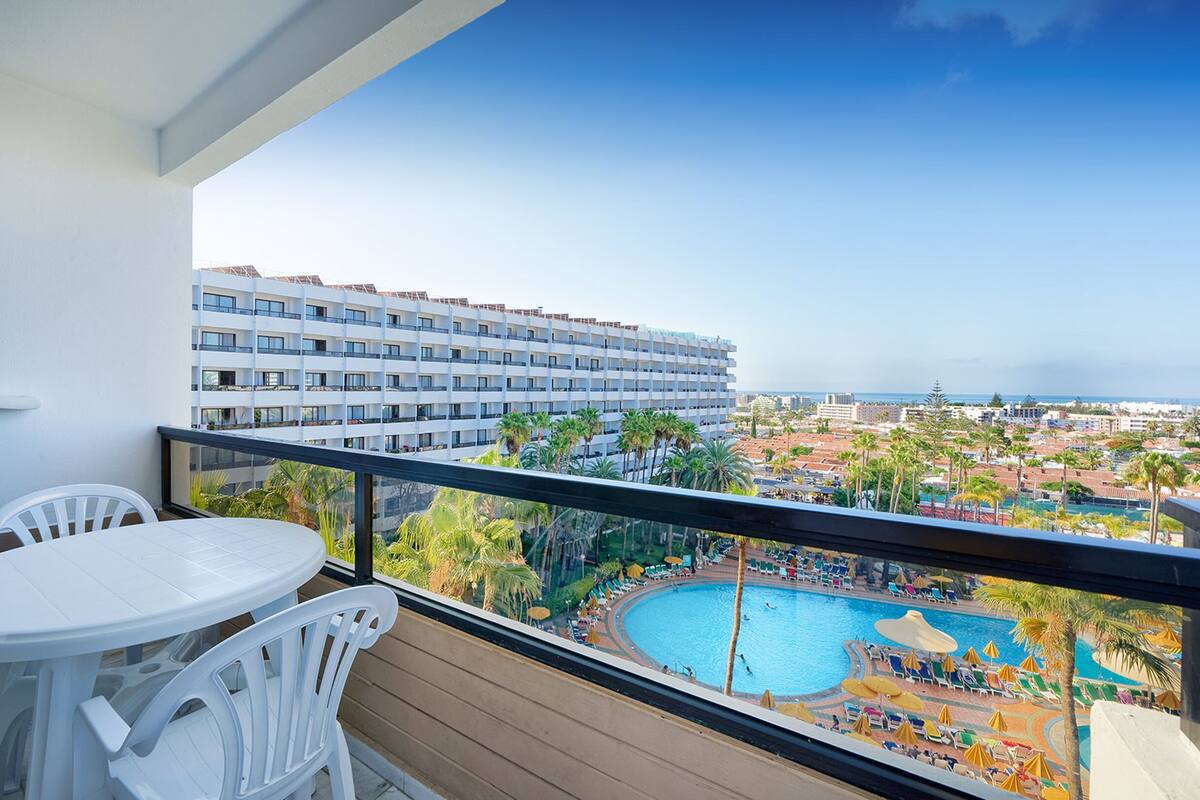 Ein Balkon mit einem weißen Tisch und Stühlen, der auf einen Poolbereich mit Sonnenschirmen und Liegestühlen sowie auf ein Hotelgebäude und das Meer blickt.