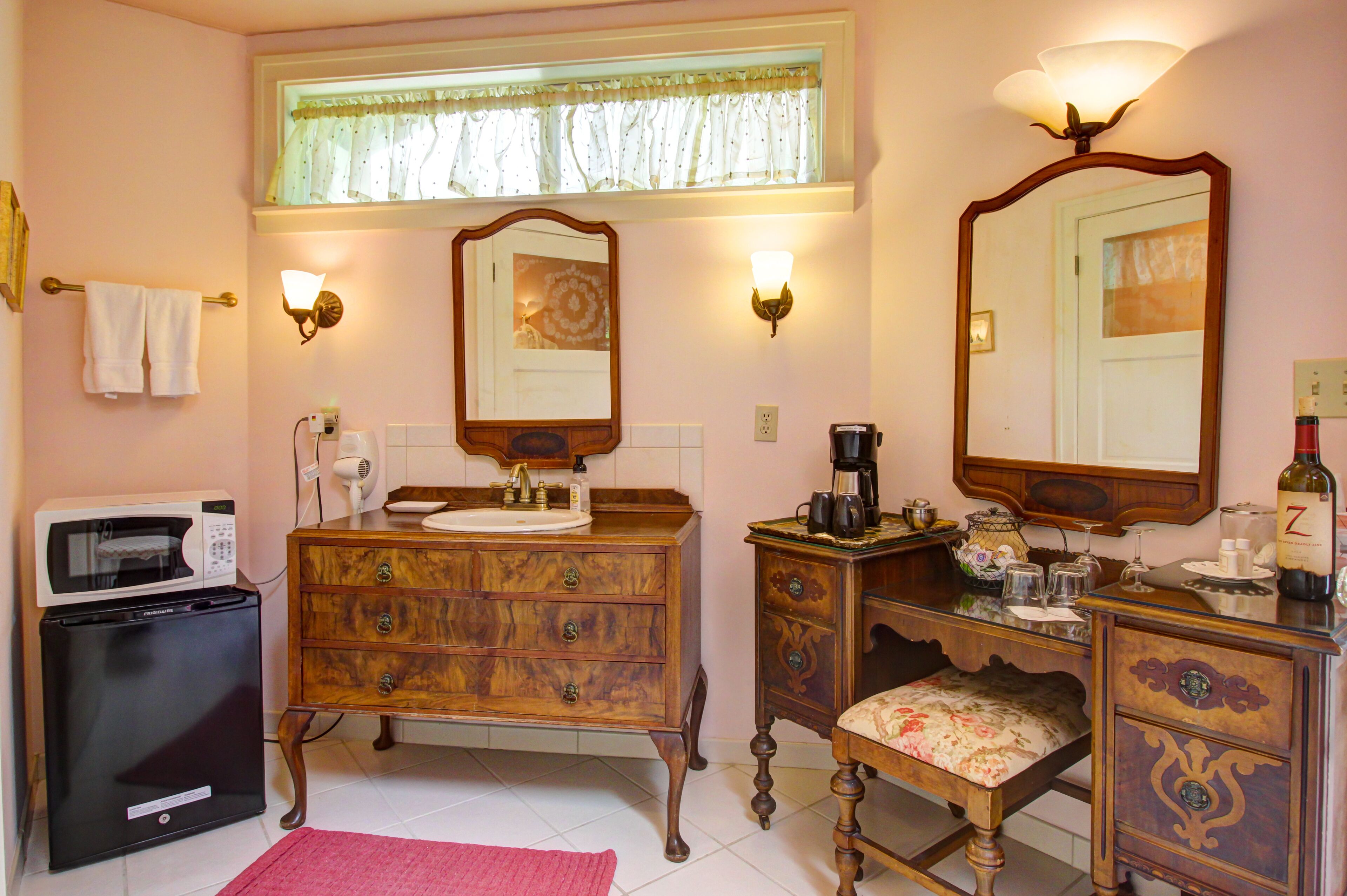 Un dormitorio pequeño con un tocador de madera, una mesita de noche, una silla, un espejo, un microondas y una ventana con cortinas.