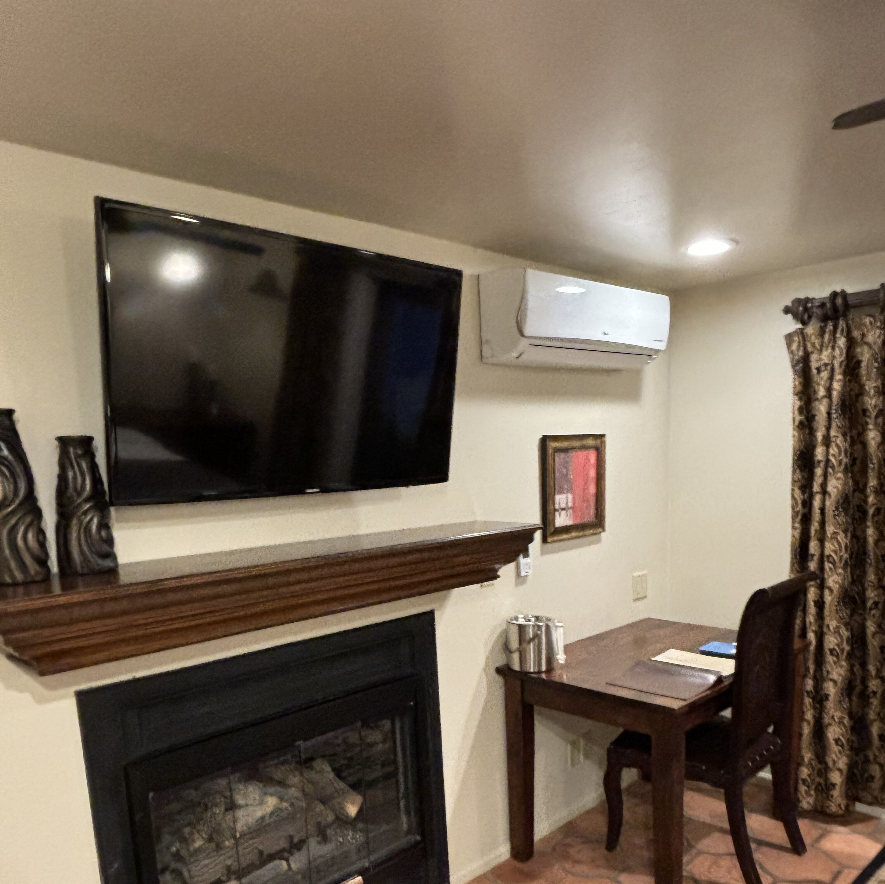 A living room with a flat-screen TV mounted above a fireplace, a table with chairs, and a patterned curtain.