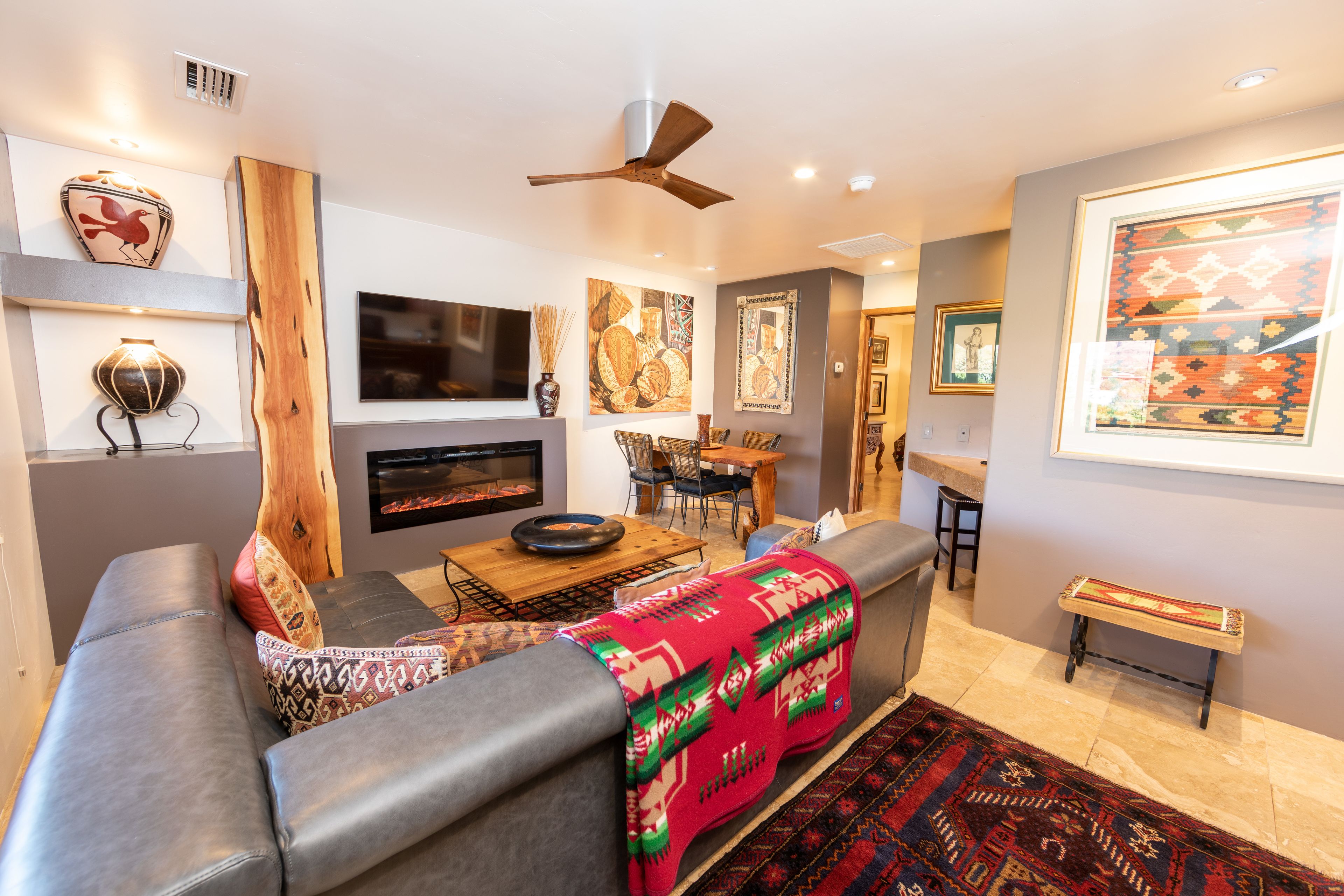 A modern living room with a gray sofa, a wooden coffee table, and a red blanket. There is a fireplace, a ceiling fan, and framed artwork on the walls.